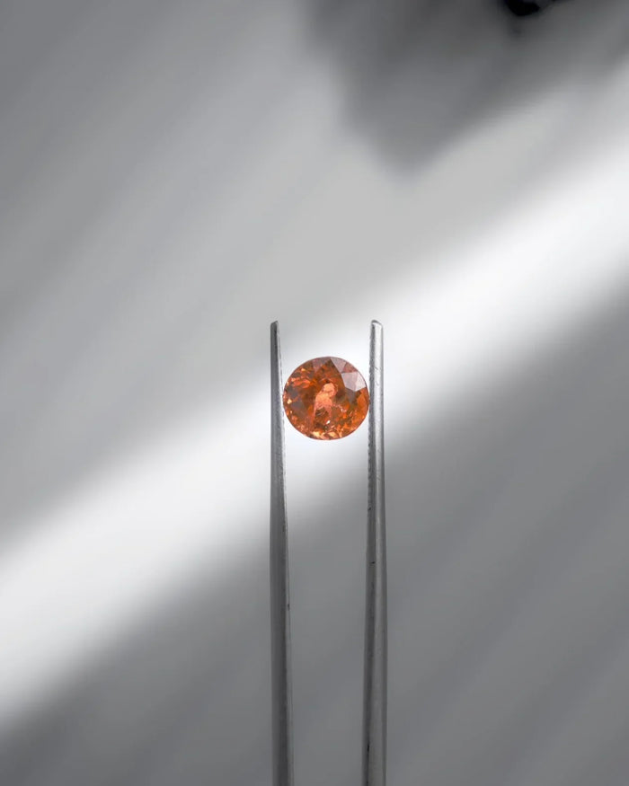 Faceted orange gemstone held by tweezers, showcasing brilliance and elegance for fine jewelry.