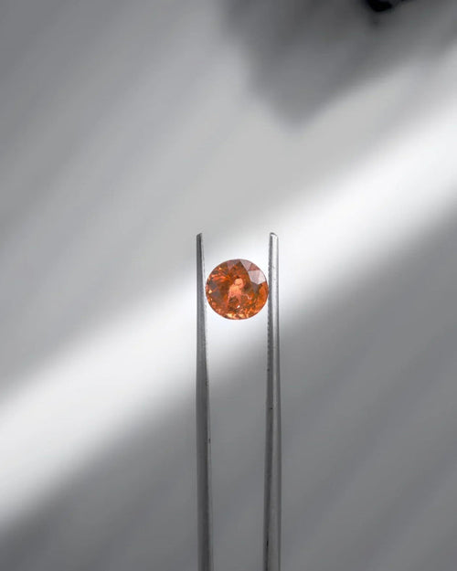 Faceted orange gemstone held by tweezers, showcasing brilliance and elegance for fine jewelry.